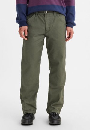 Mann trägt olivgrüne gerade geschnittene Hose, schwarze Turnschuhe und ein langärmliges Hemd mit horizontalen Streifen in Marineblau und Bordeaux.