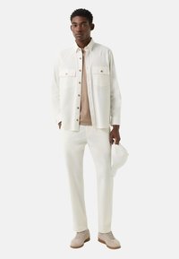 Biała koszula zapinana na guziki z kieszeniami na piersiach, jasnobeżowy t-shirt, białe spodnie i beżowe trampki. Trzyma białą czapkę.