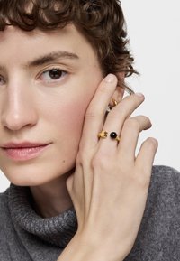 Anillos de oro con detalles negros en los dedos, llevados sobre un suéter de cuello alto gris. Primer plano de manos y rostro, resaltando texturas y detalles.