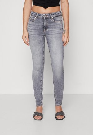 Kobieta ubrana w wyblakłe szare obcisłe dżinsy, czarny krótki top, czarne sandały na klamry oraz z małymi tatuażami na przedramionach na jednolitym tle.