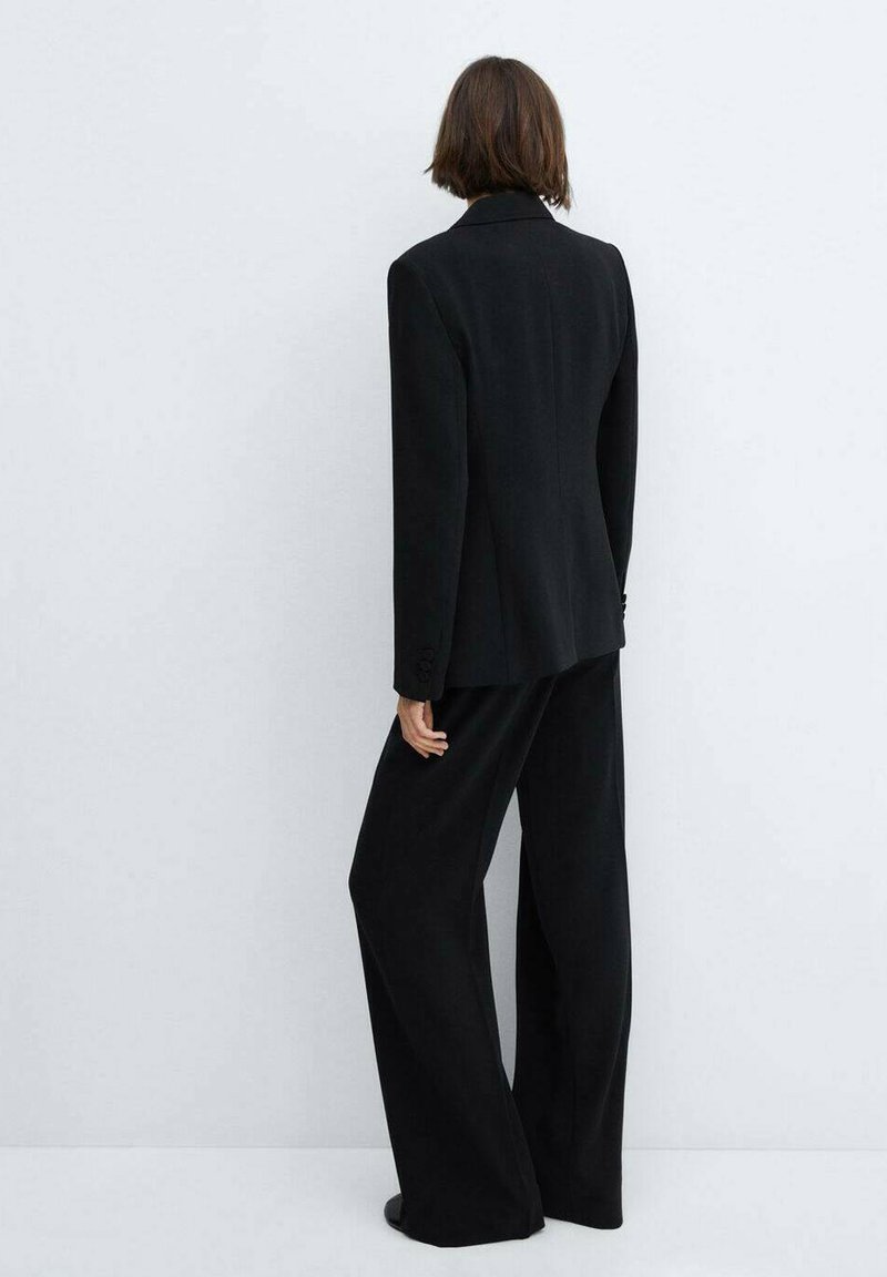 Schwarzer Anzug mit weitem Hosenbein und strukturiertem Blazer. Glattes Material und scharfe Revers mit knopfartigen Akzenten an den Ärmeln.