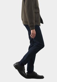 Olivenfarbener Fleece-Jacke mit einer Fronttasche, kombiniert mit marineblauen Chinos und schwarzen, schnürbaren Lederschuhen, die eine matte Textur und einen strukturierten Schnitt aufweisen.