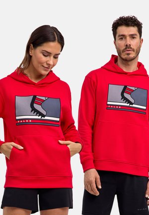 Mężczyzna i kobieta w czerwonych bluzach z kapturem z przodu z grafiką łyżwy i napisem „FALKE”, oboje ubrani są w czarne spodnie.