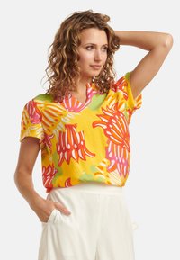 Kurzärmlige gelbe Bluse mit großen roten und rosa Blumenmustern, lockerer Passform und V-Ausschnitt. Leichte und strukturierte Stoffqualität.