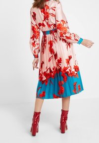 Blumenkleid in Midi-Länge mit langen Ärmeln, Druck in Rosa und Rot auf einem plissierten Stoff, mit türkisem Saum und tailliertem Schnitt. Rote Stiefeletten mit Absatz.