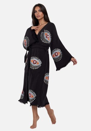 Kobieta ubrana w czarną sukienkę w stylu kimono z szerokimi rękawami i dużymi, kolorowymi wzorami oczu, stojąca boso na jednolitym tle.