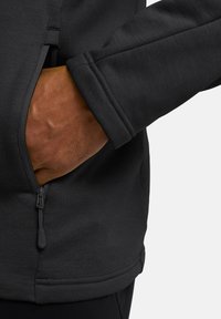Giacca di fleece nera con tasca con zip e polsini aderenti. Il tessuto è liscio, offrendo una consistenza morbida e un leggero lucido.