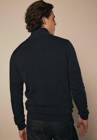 Marineblauwe gebreide trui met een hoge kraag, geribbelde manchetten en boorden, gedragen met donkere denim. De textuur lijkt zacht en aansluitend.