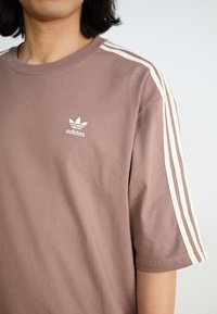 Rudas medvilninis marškinėliai su apvalia iškirpte, kuriuose yra trys baltos juostos ant pečių ir mažas baltas Adidas logotipas ant krūtinės.