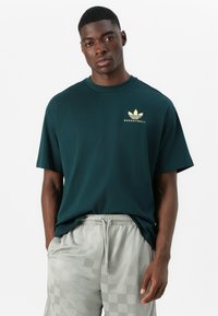 Donkergroen katoenen t-shirt met korte mouwen, voorzien van een gouden "BASKETBALL" logo en een Adidas trefoil embleem op de borst.