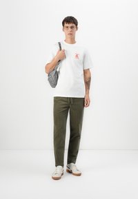 White cotton t-shirt with red graphic print, olive green corduroy pants, gray shoulder bag, and white sneakers with brown accents.