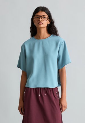 Femme aux longs cheveux foncés portant de grosses lunettes, un haut ample à manches courtes bleu clair et une jupe bordeaux avec des poches zippées.