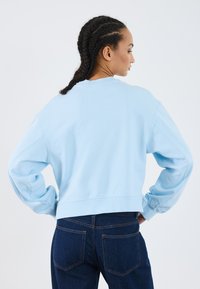 Sweatshirt azul claro, cortado, com mangas longas, gola canelada e corte descontraído, combinado com jeans de denim escuro.