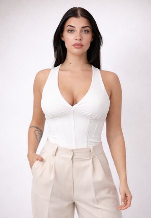 Donna con lunghi capelli scuri, indossa una canotta bianca senza maniche e pantaloni beige a vita alta, in piedi con una mano in tasca.