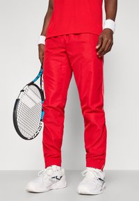 Rdeče športne hlače z belimi stranskimi črtami, elastičnimi manšetami pri gležnjih, sijoča tekstura, v kombinaciji z belimi supergami in teniškim rekvizitom.