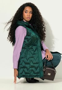 Gilet trapuntato verde con colletto alto sopra una camicia a righe viola, abbinato a pantaloni turchesi e una piccola borsa marrone. Stivaletti neri con punta visibili.