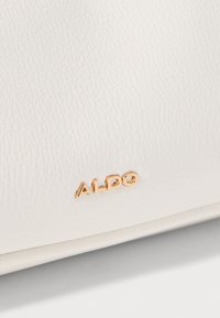 Bijela kožna torba s teksturiranom površinom, s zlatnim logotipom "ALDO". Dizajn je minimalistički s čistim linijama i bez dodatnih akcenta.