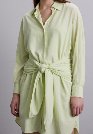 Femme portant une robe chemise manches longues vert clair avec une large ceinture en tissu nouée en nœud à la taille.