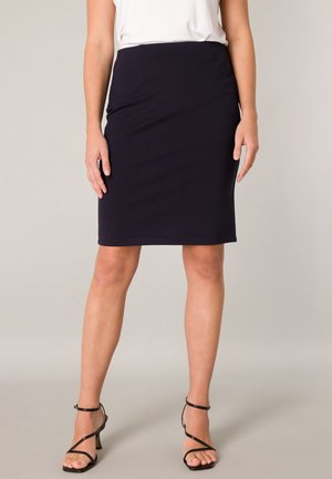 Marineblauwe kokerrok tot de knie, gemaakt van rekbare stof, met een hoge tailleband en een gladde textuur. Gecombineerd met zwarte sandaaltjes met straps.