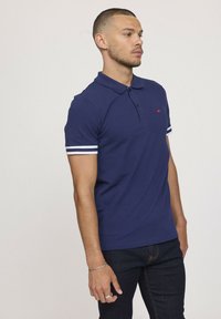 Polo shirt bleu marine en coton, avec un col, une patte de boutonnage à deux boutons, et des accents rayés blancs sur les manches courtes. Petit logo rouge sur la poitrine.