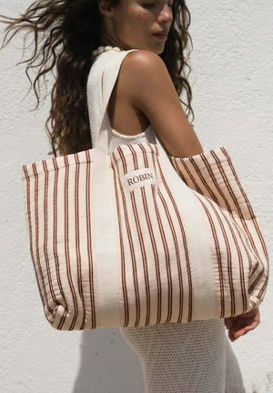 Mujer con un vestido blanco texturizado que lleva un gran bolso tote beige y marrón a rayas con la etiqueta "ROBIN" sobre su hombro frente a una pared blanca.