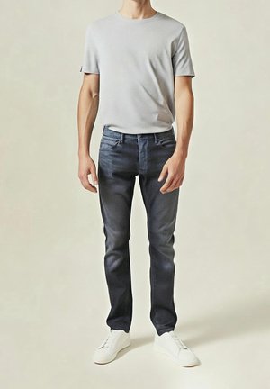 Homme portant un t-shirt gris clair, un jean slim bleu foncé et des baskets blanches, debout devant un fond uni.
