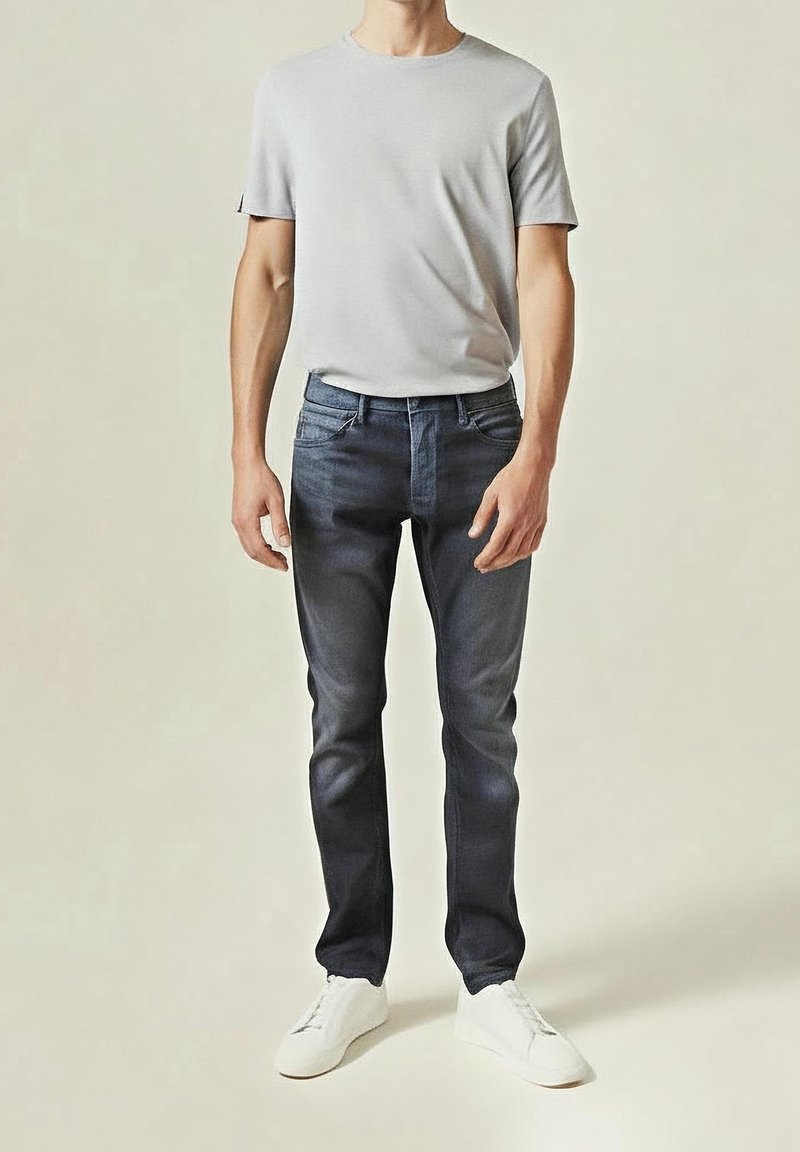 Homme portant un t-shirt gris clair, un jean slim bleu foncé et des baskets blanches, debout devant un fond uni.