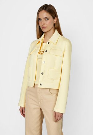 Frau trägt eine hellgelbe Jacke über einem gelben T-Shirt mit Grafik und beigefarbenen Hosen, steht vor einem schlichten weißen Hintergrund.