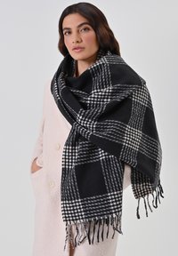 Zwart-wit geruite wollen sjaal met franjes, over een lichtroze jas gedrapeerd. Het patroon heeft dikke en dunne lijnen in een klassiek ontwerp.