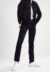 Quiltad svart bomberjacka med hög krage, som kombineras med mörkblå åtsittande jeans och vita sneakers med röda accenter.