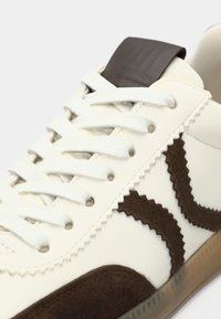 Valge nahast ja seemisnahast toss, millel on pruunid siksakmustrilised detailid, tasased paelad ja tekstuuriga pruun kontsaluuk.