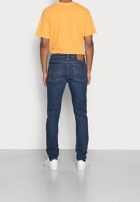 Hombre con una camiseta naranja, jeans de mezclilla azules y zapatillas blancas, de pie mirando hacia atrás contra un fondo blanco liso.