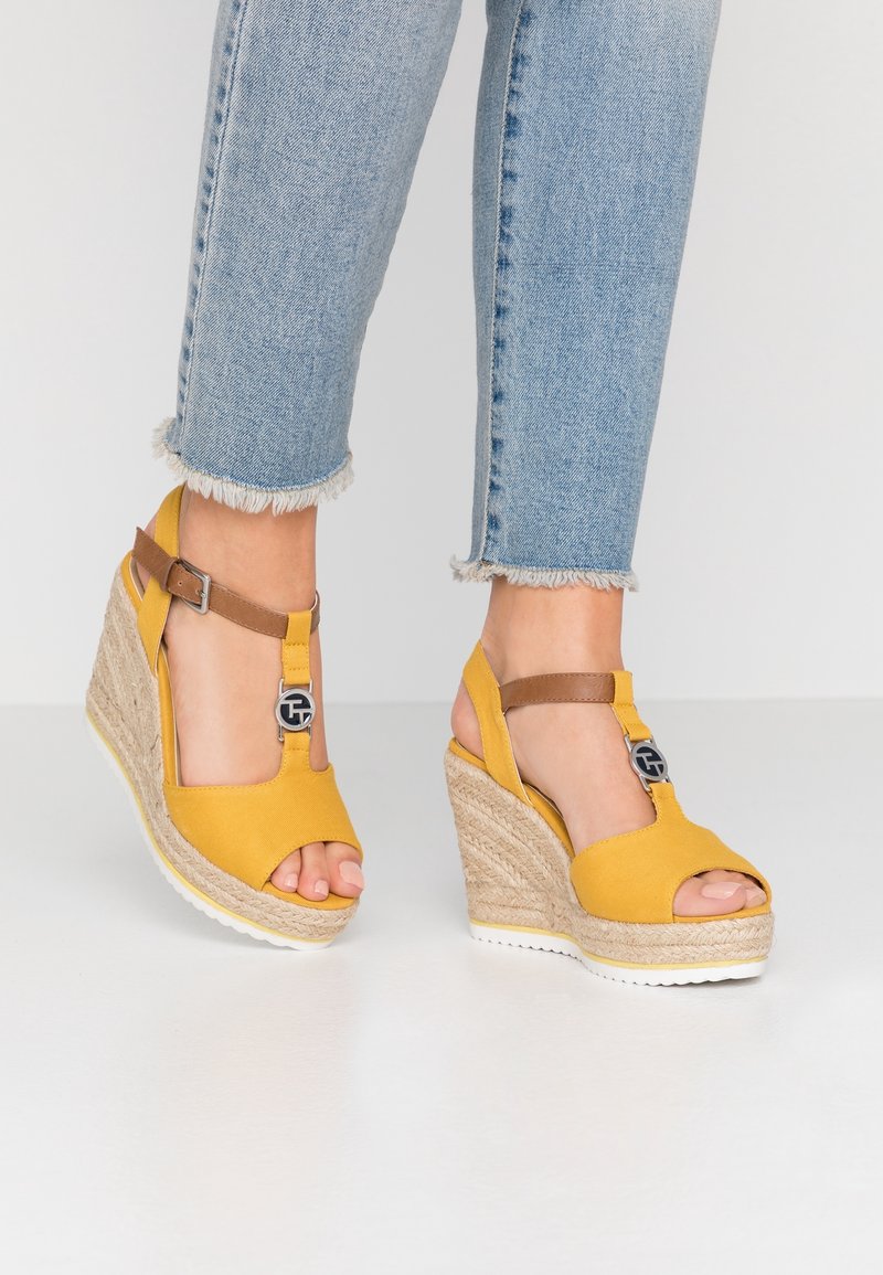 Pieds portant des sandales espadrilles compensées jaunes avec des lanières marron autour de la cheville, associées à un jean bleu clair à ourlet effiloché sur un sol blanc.