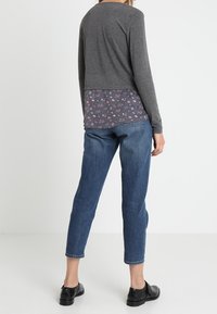 Grå langærmet trøje lagd over en blomstret bluse, parret med blå, tapered jeans og sorte sko. Glatte teksturer og afslappet pasform.