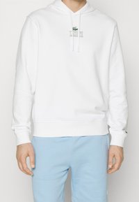 Vit hoodie med känguruficka, logotyp fram, dragsko på huvan och ribbstickade muddar, kombinerad med ljusblå mjukisbyxor.