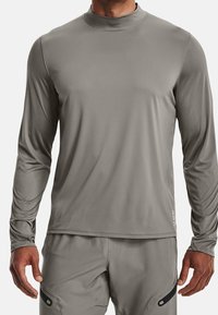 Mann in einem enganliegenden, langärmeligen grauen Sportshirt und passenden Hosen mit schwarzen Reißverschlussdetails an den Oberschenkeln.