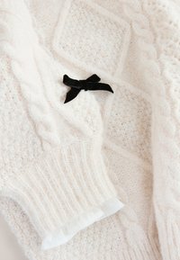 Suéter de punto color crema con patrones de trenzado y un panel texturizado, con un lazo de terciopelo negro como detalle y puños suaves con volantes.