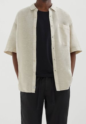 Hombre con camisa beige de manga corta desabotonada sobre camiseta negra y pantalones negros con cuerda frente a un fondo claro y liso.