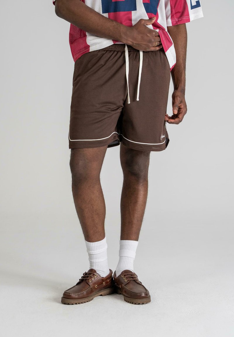 Shorts en maille marron avec des garnitures blanches et un cordon de serrage. Portés avec des chaussettes blanches et des chaussures marron. Le design comprend des fentes latérales pour plus de confort.