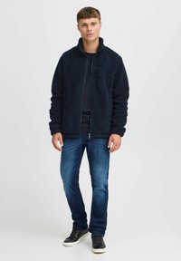 Veste en polaire marine avec fermeture éclair à l'avant, col montant et deux poches extérieures, associée à un jean sombre et des baskets noires. Texture lisse et douce.
