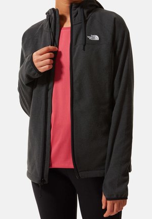 Osoba nosząca czarną bluzę polarową The North Face z zamkiem błyskawicznym na czerwonej koszulce, regulująca zamek kurtki jedną ręką.