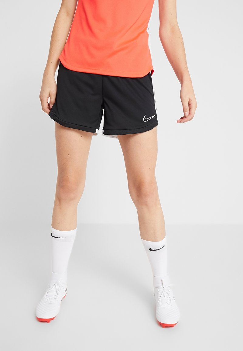 Athlète portant un short Nike noir, des chaussettes blanches Nike, des crampons blancs et un haut orange, debout devant un fond clair uni.