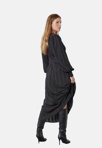 Čierne šaty s dlhým rukávom z texturovanej tkaniny, s plisovaním a bočným rozparkom, skombinované s lesklými čiernymi čižmami po kolená.