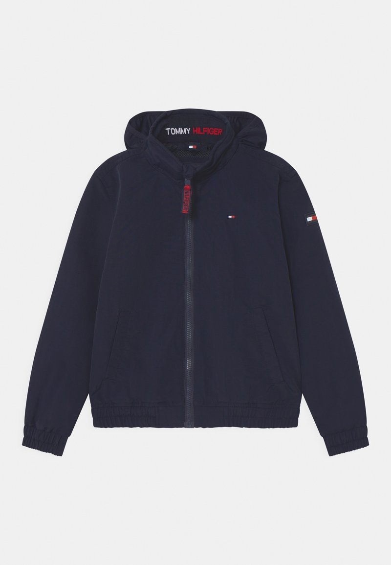 Marineblauwe windbreakerjack met een capuchon, elastische manchetten, twee frontzakken en een rode rits. Heeft een logo op de bovenkant van de linkerarm.