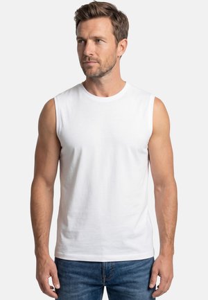 Mand med kort brunt hår iført en hvid ærmeløs skjorte og blå jeans, der kigger let mod venstre mod en ensartet lys baggrund.