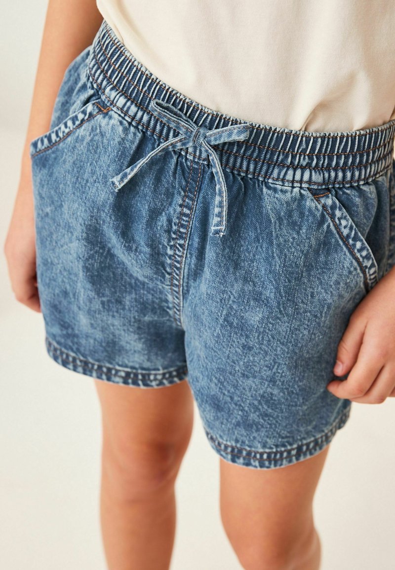 Enfant portant un short en denim bleu clair avec taille élastique et cordon, associé à une chemise blanche rentrée.