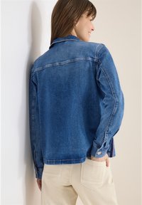 Femme portant une veste en jean bleue et un pantalon beige, debout de profil avec la main dans la poche arrière, sur un fond clair.