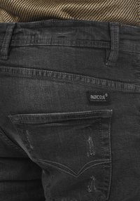 Jeans de mezclilla negra con una textura desgastada, que presentan un diseño de bolsillo trasero y una etiqueta de la marca "INDICODE" cosida en la cinturilla.