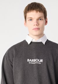 Šedá mikina s dlouhými rukávy, na přední straně s bílým logem "BARBOUR INTERNATIONAL". Nositá přes bílou košilovou halenku.
