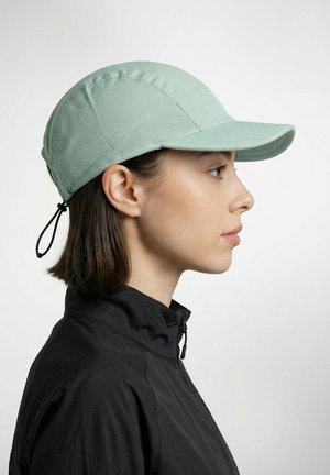 Femme en veste noire portant une casquette vert clair avec une sangle ajustable, vue de profil droit sur un fond uni.